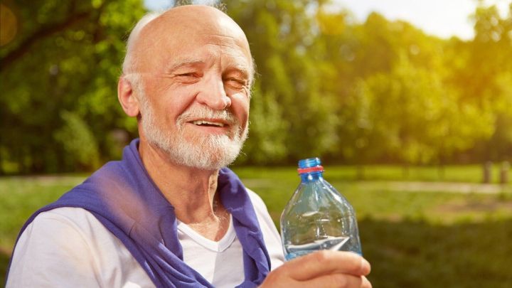 Ein &auml;lterer Mann trinkt im Sommer drau&szlig;en eine Wasserflasche, w&auml;hrend er l&auml;chelnd in einem sonnigen Park sitzt.