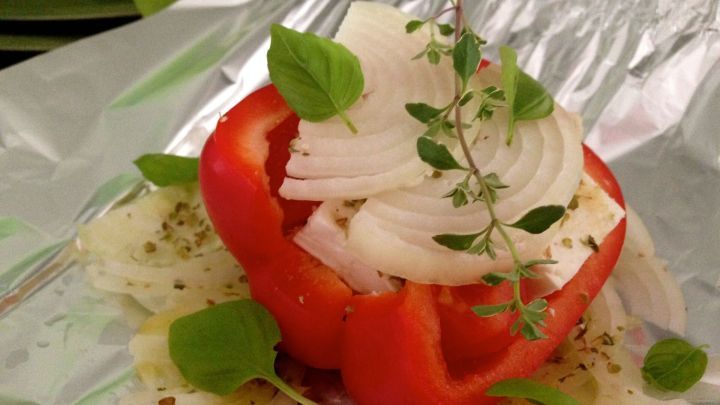 Eine gef&uuml;llte Paprikah&auml;lfte mit Ziegenk&auml;se ist in Alufolie platziert, garniert mit Zwiebeln und Kr&auml;utern.
