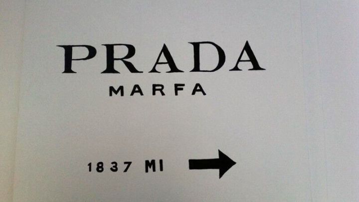 Ein Schild mit der Aufschrift &bdquo;PRADA MARFA&ldquo; zeigt, dass man 1837 Meilen in diese Richtung f&auml;hrt, auf wei&szlig;em Hintergrund.