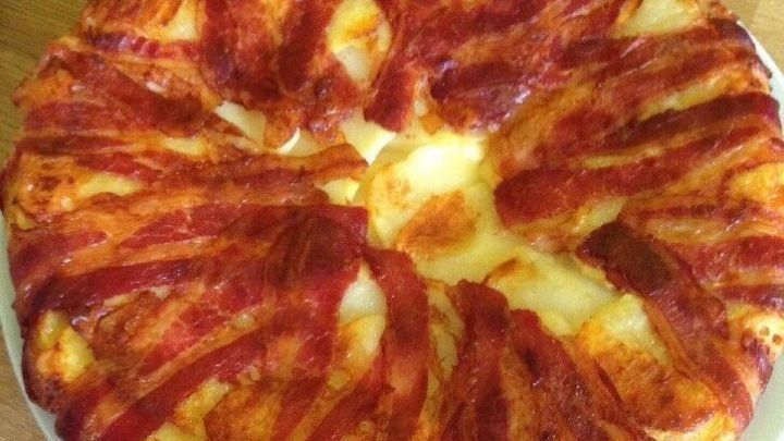 Ein runder Kartoffel-Bacon-Kuchen liegt auf einem Holzbrett und zeigt knusprigen Bacon, der attraktiv angeordnet ist.