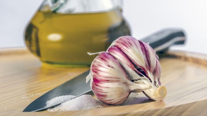 Frischer Knoblauch liegt auf einem Holzbrett neben einem scharfen Messer und einem Glas mit Oliven&ouml;l im Hintergrund.