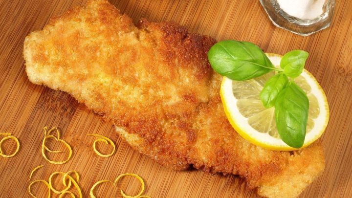Ein gebackenes Putenschnitzel liegt auf einem Holzbrett, garniert mit Zitrone und Basilikum, umgeben von weiteren Zutaten.
