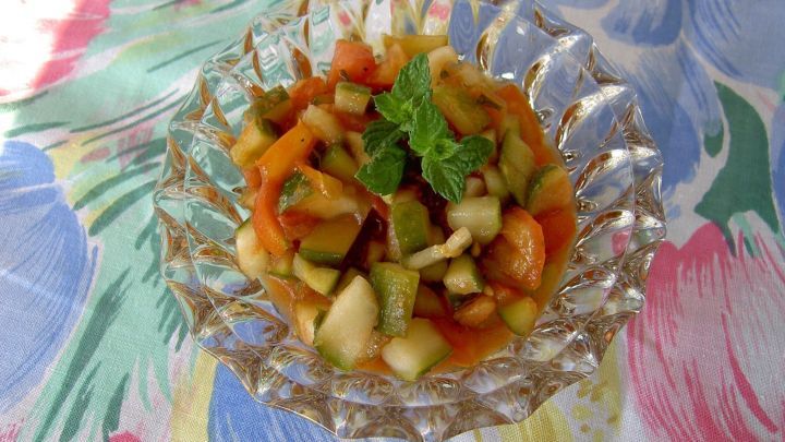 Ein Kristallsch&auml;lchen h&auml;lt bunte Salsa-Salatw&uuml;rfel, garniert mit Minzbl&auml;ttern, auf einem Tisch mit floralem Tischtuch.