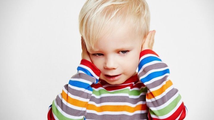 Ein Junge mit einer gestreiften Bluse h&auml;lt sich die Ohren zu und zeigt Symptome einer Mittelohrentz&uuml;ndung in einem neutralen Raum.