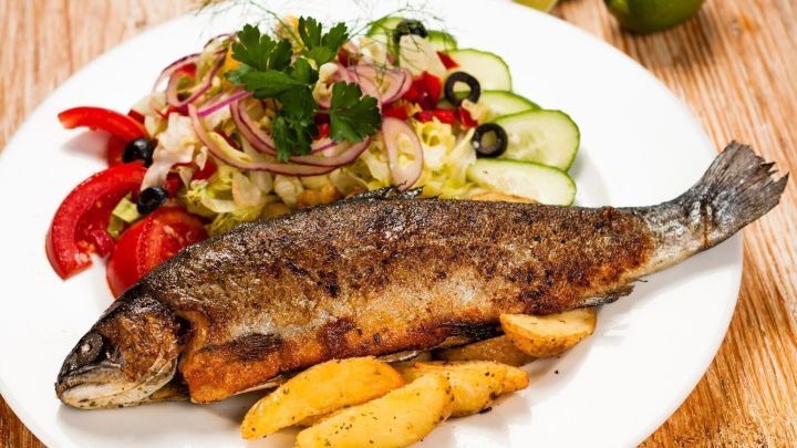 Eine panierte Forelle M&uuml;llerin liegt auf einem Teller, umgeben von frischem Salat und knusprigen Kartoffelecken.