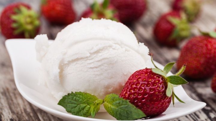 Eine Schale mit cremigem Eis liegt auf einem rustikalen Tisch, umgeben von frischen Erdbeeren und Minzbl&auml;ttern.