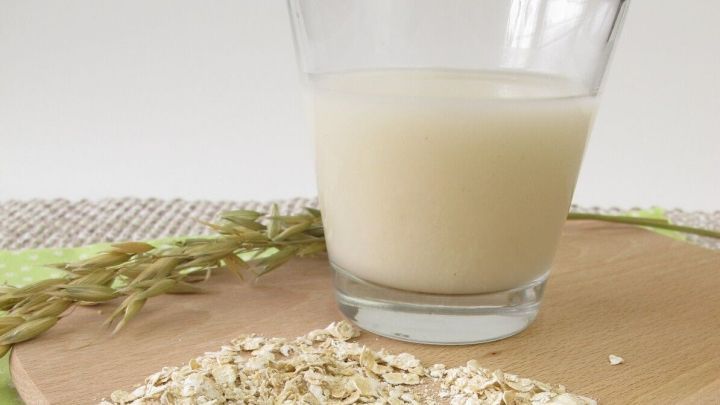 Ein Glas Hafermilch steht auf einem Holzbrett, umgeben von Haferflocken, auf einem Textiluntergrund.