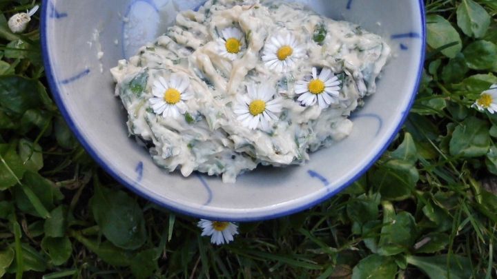 Eine Schale gef&uuml;llt mit Kr&auml;uterbutter, dekoriert mit G&auml;nsebl&uuml;mchen, steht auf einer Wiese umgeben von frischem Gr&uuml;n.