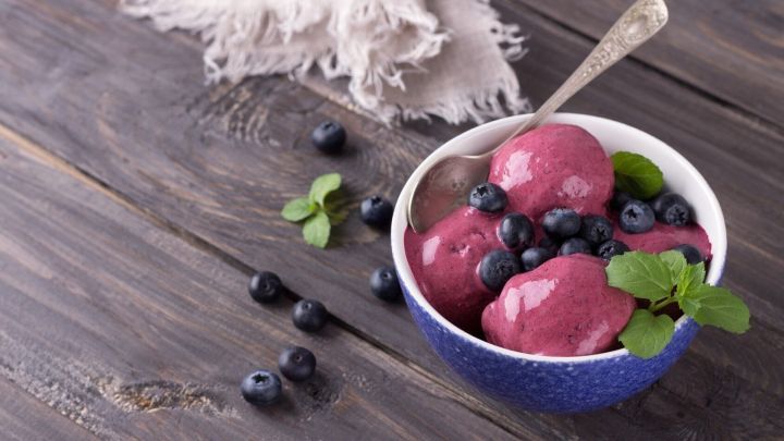 In einer Sch&uuml;ssel liegt fruchtiges Eis, garniert mit frischen Heidelbeeren und Minzbl&auml;ttern, auf einem rustikalen Holz Tisch.