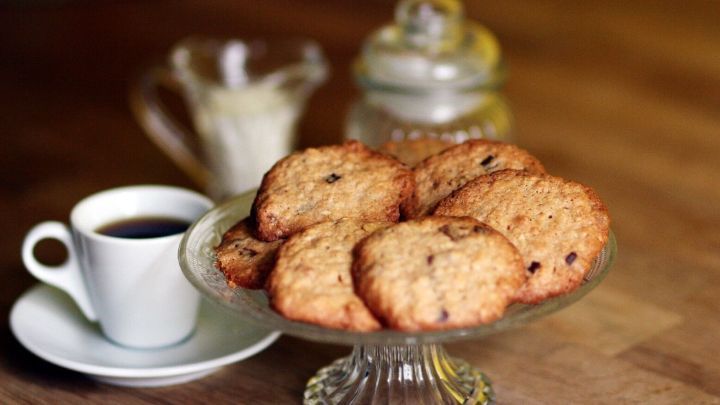 Ein Teller mit leckeren Cookies aus M&uuml;sliresten steht neben einer Tasse Kaffee auf einem Holztisch, gem&uuml;tliche Atmosph&auml;re.