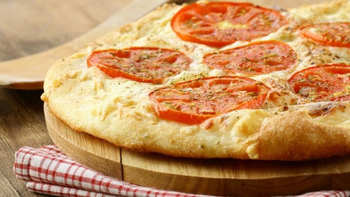 Eine Sommerpizza mit Tomatenscheiben liegt auf einem Holzbrett vor einer rot-wei&szlig; karierten Serviette.