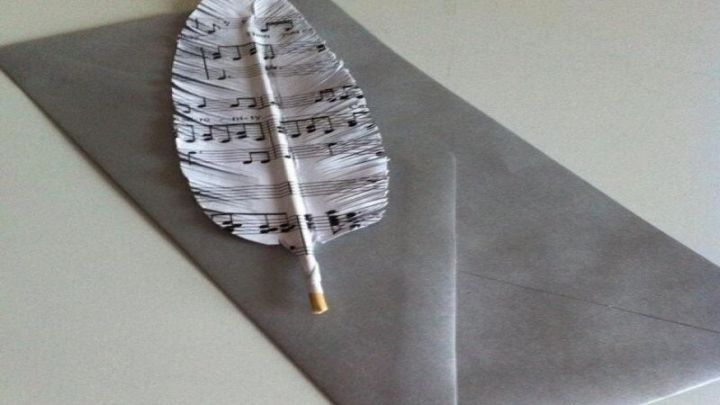 Eine Deko-Feder aus Papier liegt auf einem grauen Umschlag, verziert mit Notenlinien, die eine kreative Note verleihen.