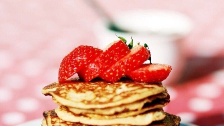 Auf einem Teller befinden sich stapelweise goldbraune Bananen-Pancakes, dekoriert mit frischen Erdbeeren. Im Hintergrund ist eine rosa, gepunktete Tischdecke zu sehen.