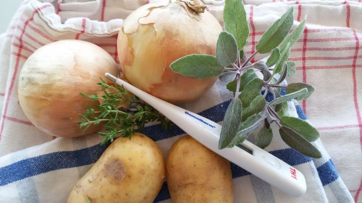 Zwei Zwiebeln und Kartoffeln liegen auf einem K&uuml;chentuch, umgeben von Kr&auml;utern, bereit f&uuml;r alte Hausmittel bei Beschwerden.