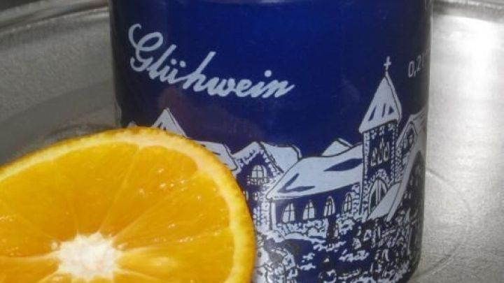 Blaue Tasse mit der Aufschrift "Gl&uuml;hwein" zeigt eine Winterlandschaft; daneben liegt eine halbierte Orange auf einem Tisch.