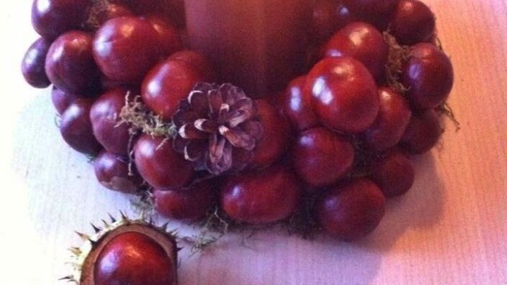 Ein Kastanien-Kerzenkranz aus gl&auml;nzenden Kastanien umgibt eine brennende Kerze und steht auf einem hellen Untergrund.