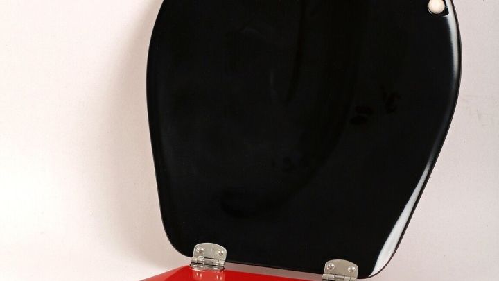 Ein rot lackierter Toilettensitz mit einem schwarzen Deckel steht ge&ouml;ffnet. Er zeigt einen modernen, farbenfrohen Stil in einem neutralen Raum.