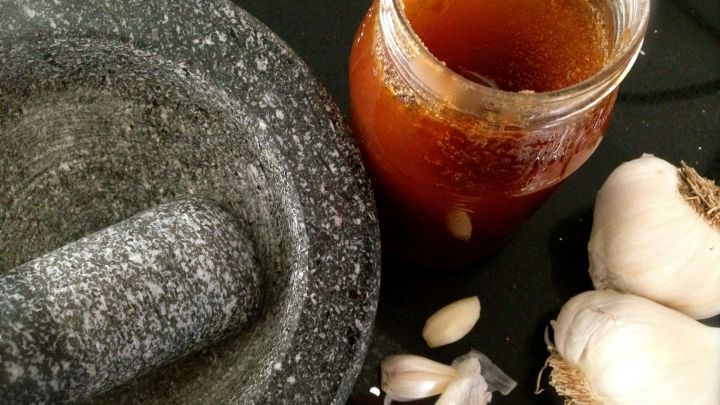 Ein M&ouml;rser liegt neben einem Glas Knoblauch-Honig-Paste und frischen Knoblauchzehen auf einem dunklen Untergrund.