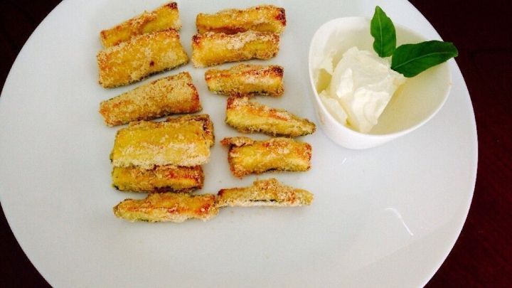 Zucchini-Sticks ruhen knusprig auf einem wei&szlig;en Teller mit einem Becher Dip, verziert mit einem gr&uuml;nen Kr&auml;uterblatt.
