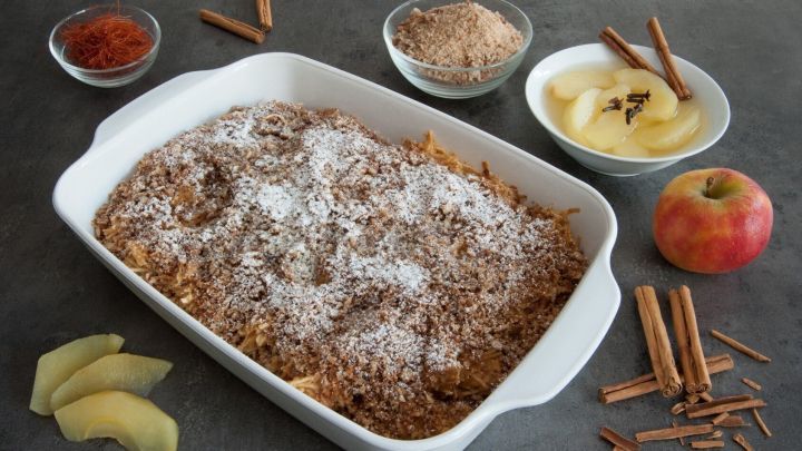 Ein Apfelauflauf mit Roggenbrot ist goldbraun gebacken, bestreut mit Puderzucker, umgeben von &Auml;pfeln und Gew&uuml;rzen.