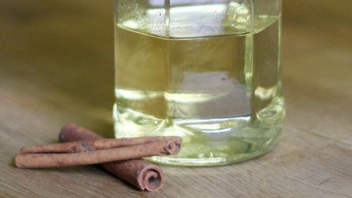 Eine Glasflasche mit Zimt&ouml;l steht auf einem Holztisch, umgeben von Zimtstangen vor gr&uuml;nem Hintergrund.