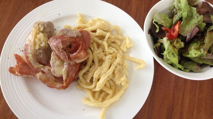 Zartes Schweinefilet im Speckmantel liegt auf einem wei&szlig;en Teller, umgeben von Sp&auml;tzle und einem frischen Salat.