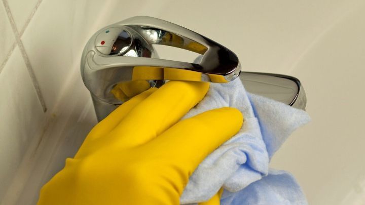 Ein Hand mit einem gelben Handschuh wischt einen Wasserhahn mit einem blauen Tuch, um l&auml;stige Wasserflecken zu entfernen.