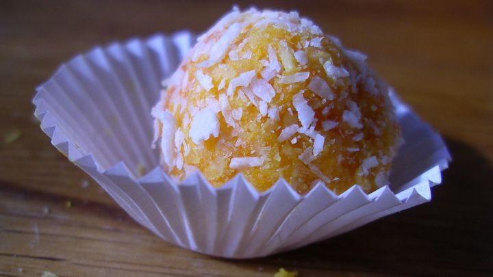 Eine orangefarbene Mandel-Kokos-Praline liegt in einer Papierkapsel und funkelt appetitlich auf einem Holzuntergrund.