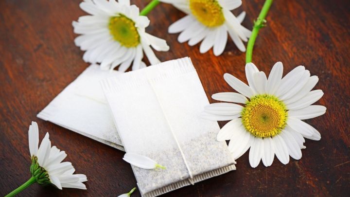 Kamillentee-Beutel liegen auf einem Holztisch, umgeben von frischen Kamillenbl&uuml;ten, die eine beruhigende Wirkung andeuten.