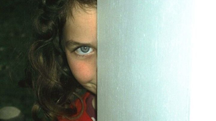 Ein M&auml;dchen mit lockigem Haar blickt neugierig hinter einer S&auml;ule hervor, w&auml;hrend der Hintergrund unscharf bleibt.