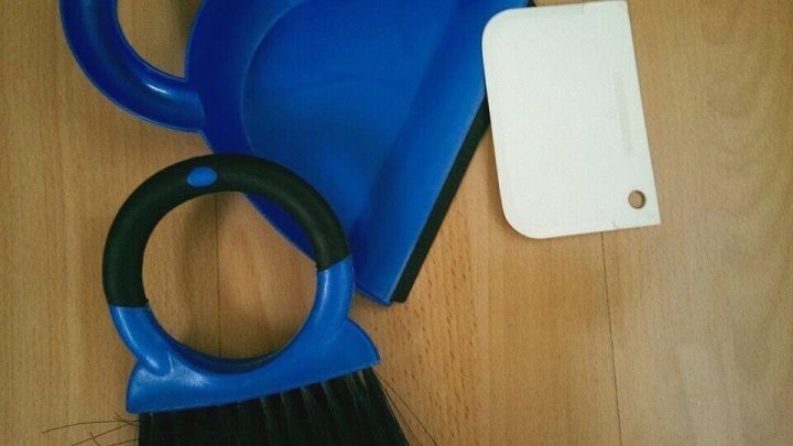 Ein blauer Besen mit Schaufel liegt auf einem Holzboden, bereit, ein rohes Ei aufzunehmen. Die Umgebung wirkt sauber und ordentlich.