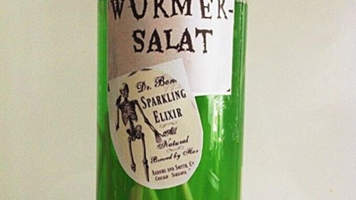 Ein Glas mit gr&uuml;nem &bdquo;W&uuml;rmersalat&ldquo; steht auf einem Tisch, gef&uuml;llt mit langen, gr&uuml;nen Essenzweigen, passend zu Halloween.