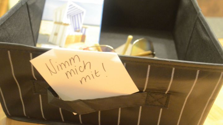 Ein schwarzer Aufbewahrungskorb steht auf einem Tisch. Ein Zettel mit der Aufschrift &bdquo;Nimm mich mit!&ldquo; ist sichtbar.