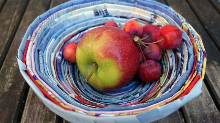 Ein bunter Papierschale h&auml;lt einen roten Apfel und Kirschen auf einem Holztisch im Freien.