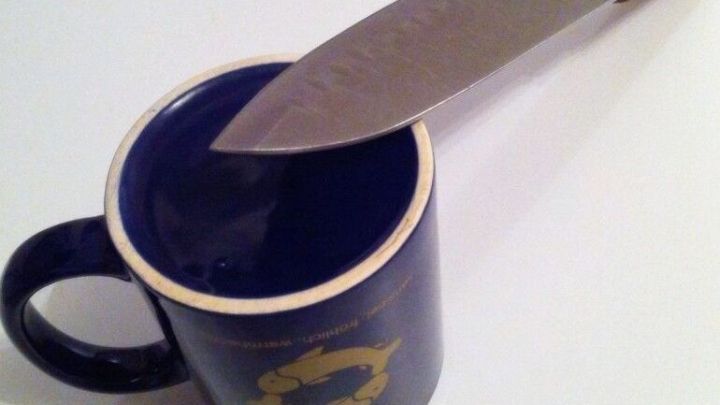 Ein scharfes Messer liegt auf dem Rand einer blauen Tasse, die auf einer wei&szlig;en Oberfl&auml;che steht.