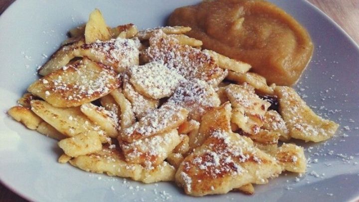 Fluffiger Kaiserschmarrn liegt auf einem wei&szlig;en Teller, wird mit Puderzucker bestreut und hat eine Beilage aus Apfelmus.