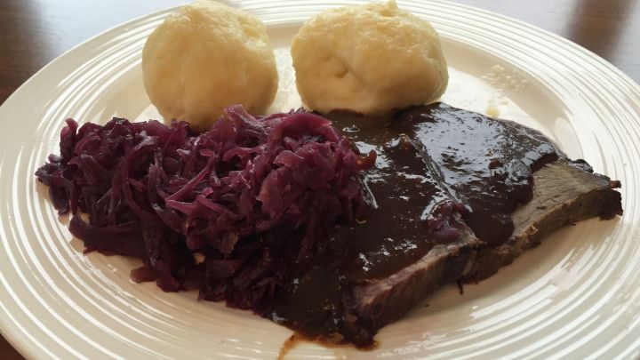 Kartoffelkn&ouml;del liegen neben einem saftigen St&uuml;ck Fleisch und Rotkohl auf einem wei&szlig;en Teller in einer gem&uuml;tlichen Umgebung.