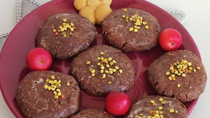 Kleine Schokopl&auml;tzchen mit Hirse und N&uuml;ssen liegen auf einem roten Teller, umgeben von kleinen roten &Auml;pfeln und einem Keksb&auml;rchen.