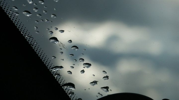 Wassertropfen liegen klar auf der Autoscheibe, w&auml;hrend graue Wolken im Hintergrund sichtbar sind, die eine nasse Umgebung andeuten.
