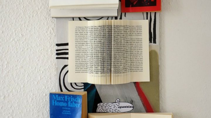Ein aufrecht h&auml;ngendes Kunstwerk zeigt alte B&uuml;cher, die kreativ angeordnet sind, mit Seiten, die aufgeschlagen sind, vor einem minimalistischen Hintergrund.