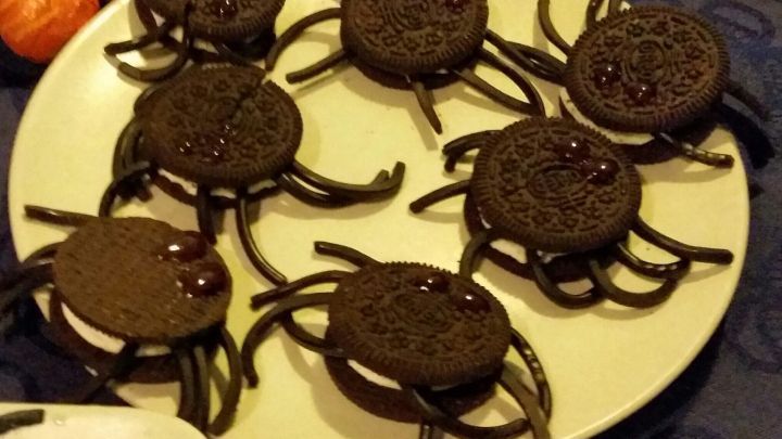 Sechs schaurige Spinnenkekse liegen auf einem Teller, umgeben von weiteren Snacks auf einem dunklen Tisch.