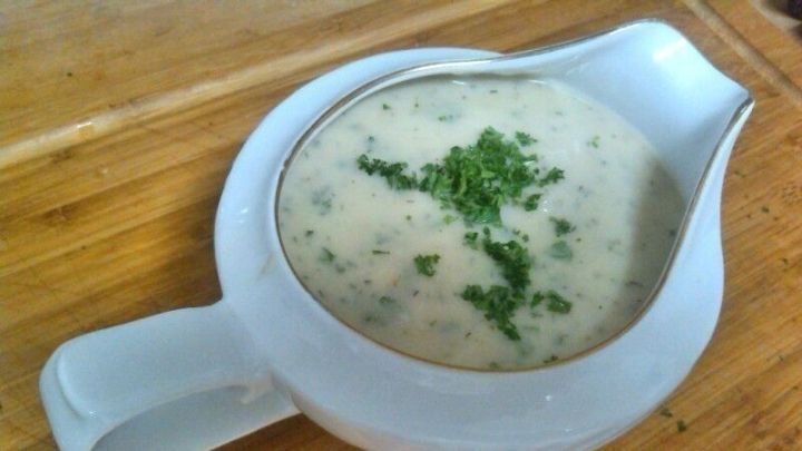 Eine wei&szlig;e Gorgonzolaso&szlig;e mit frischer Petersilie wird in einer eleganten Sauciere auf einem Holzbrett serviert.