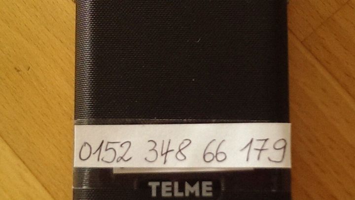 Ein schwarzes Handy liegt auf einem Holztisch, mit einer Aufschrift f&uuml;r wichtige Telefonnummern auf der R&uuml;ckseite.