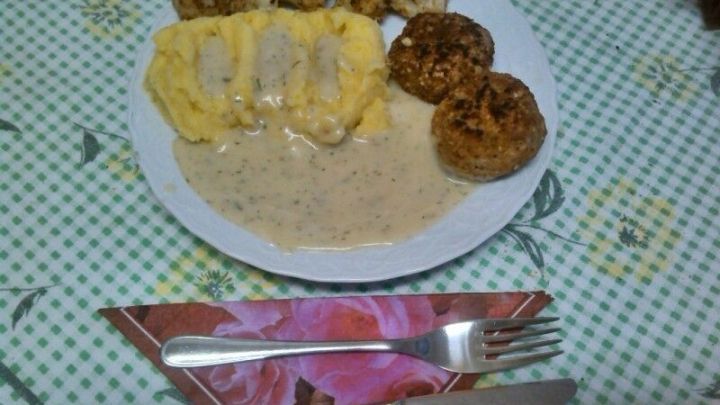 Bouletten aus Blumenkohl liegen neben cremigem P&uuml;ree und Sauce auf einem wei&szlig;en Teller, umgeben von einem gemusterte Tischdecke.