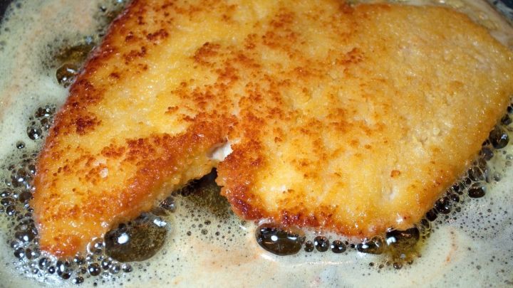 Ein goldbraun paniertes Schnitzel br&auml;t in einer Pfanne mit sprudelndem Fett, umgeben von blubbernden Blasen.
