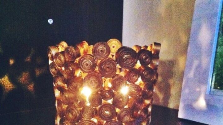 Ein Kerzenhalter aus Altpapier steht auf einem Regal und beleuchtet den Raum mit warmem Licht, umgeben von dunklem Hintergrund.