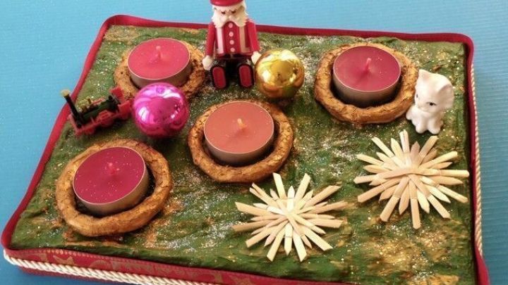 Ein rechteckiger Adventskranz mit vier Kerzen, goldenen und bunten Kugeln sowie weihnachtlichen Figuren auf gr&uuml;nem Grund.