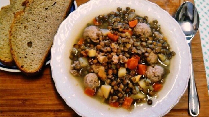Eine Sch&uuml;ssel Linsensuppe mit Hackfleischkl&ouml;&szlig;chen und Gem&uuml;se steht auf einem Holzbrett neben zwei Scheiben Brot, auf einem bunten Tisch.