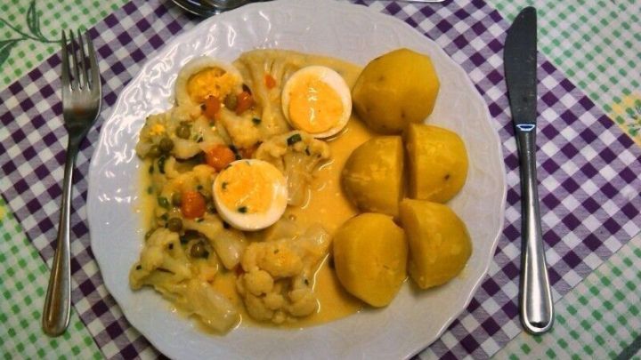 Ein Teller Blumenkohlragout mit Eieinlage und Kartoffeln liegt auf einem karierten Tischdecken, umgeben von Besteck.