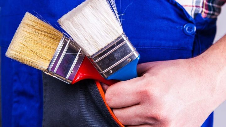 Pinsel werden von einer Hand in blauer Arbeitskleidung gehalten. Sie stecken in einer Tasche einer Jacke mit Karomuster-Hemd.